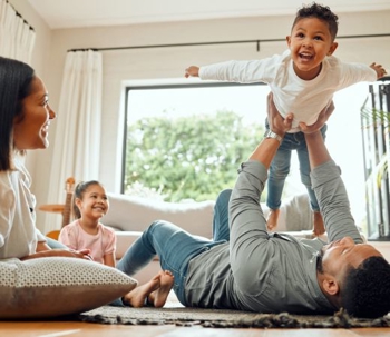 A happy family enjoys time together in a bright living room. A dad lifts his laughing son in the air as a mom and daughter watch cheerfully.