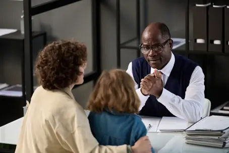 A man with glasses sits at a desk, speaking to a woman embracing a child. The woman's back is to the viewer, and the child looks towards the man.