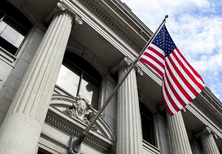 An American flag flies prominently from a pole in front of a classical building with large stone columns. The ornate, light-colored architecture suggests a government or institutional setting against a cloudy sky.